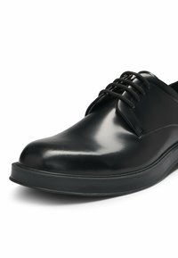 Black leather shoe with a glossy finish, rounded toe, and black laces. Features a minimalistic design and a flat rubber sole.