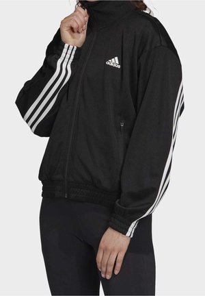 Černá zipová bunda s vysokým límcem. Obsahuje bílé logo Adidas a tři bílé pruhy na ramenou. Texturovaná tkanina, elastické manžety.