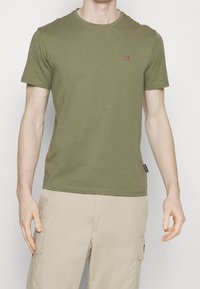 Olivengrøn bomulds t-shirt med rund hals, korte ærmer og et lille rødt og hvidt broderet emblem på brystet.