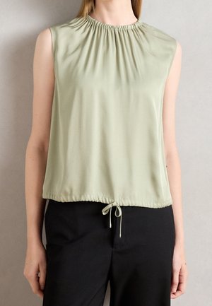 Blouse sans manches vert clair avec encolure froncée et bas à cordon ajustable portée avec un pantalon noir sur une personne.