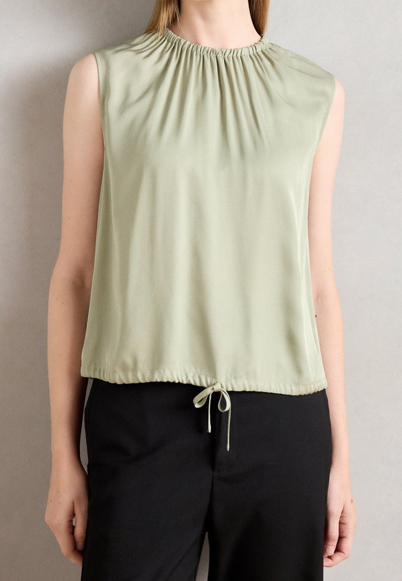 Blouse sans manches vert clair avec encolure froncée et bas à cordon ajustable portée avec un pantalon noir sur une personne.