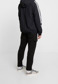 Svart huva jacka med vita ränder på ärmarna, i kombination med svarta cargobyxor och grå sneakers med vita detaljer och snören.