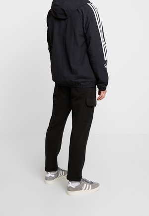Zwarte hooded jacket met witte strepen op de mouwen, gecombineerd met zwarte cargo pants en grijze sneakers met witte accenten en veters.