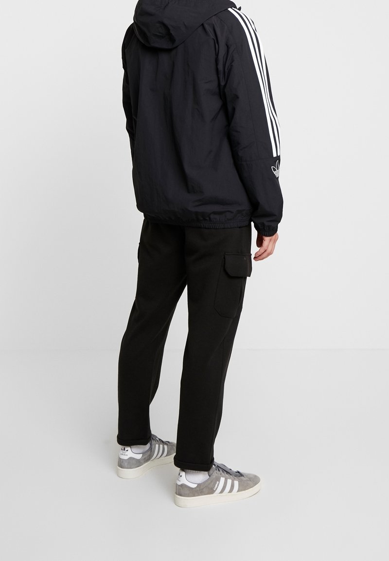 Svart huva jacka med vita ränder på ärmarna, i kombination med svarta cargobyxor och grå sneakers med vita detaljer och snören.