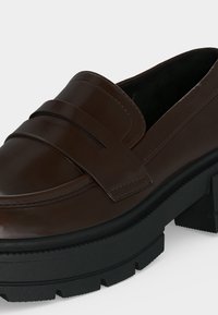 Mocassin en cuir marron foncé avec un large détail de bride et une semelle épaisse en caoutchouc noir, présenté en gros plan sur un fond clair.