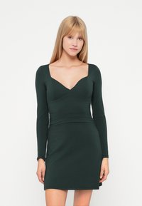 Tumma vihreä istuva mekko pitkillä hihoilla, syvän neliön muotoisella kaula-aukolla ja teksturoidulla kankaalla. Siinä on kapea siluetti ja polvipituinen helma.