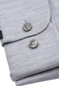 Hellgrauer Stoff mit einem subtilen Diagonal-Muster, ausgestattet mit zwei runden grauen Knöpfen und sorgfältigen Nähten. Akzente bilden ein gefaltetes Design.