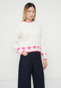 Jauna sieviete ar taisnām matēm valkā baltu adītu džemperi ar rozā sirdīm un tumšiem plašiem biksēm, stāvot uz viendabīga fona.