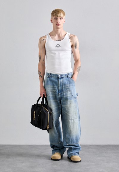 Jeune homme avec des tatouages portant un débardeur blanc, un jean loose délavé, des chaussures beiges, tenant un sac noir Moschino contre un fond gris uni.