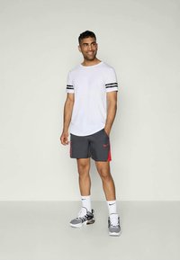 Balta sportinė marškinėliai su juodomis juostelėmis ant rankovių, derinami su pilkomis šortais, turinčiais raudonus akcentus ir „Nike“ logotipu. Pilki sportbačiai su baltais akcentais.