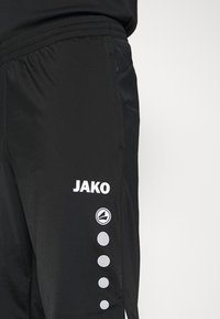 Pantalons de sport noirs avec une taille élastique, logo "JAKO" blanc et cinq accents circulaires sur le côté. Texture de tissu lisse.