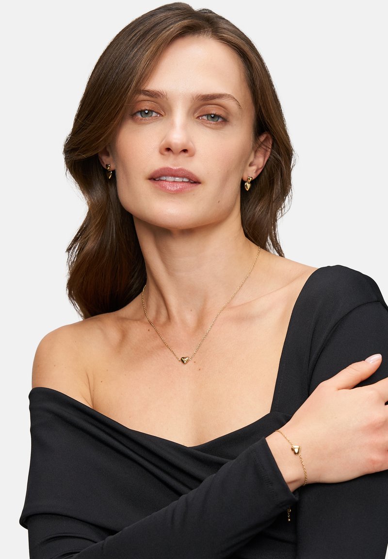 Gouden ketting en armband set met hartvormige bedels; gedragen over een off-shoulder zwarte top; gladde textuur, minimalistisch ontwerp.