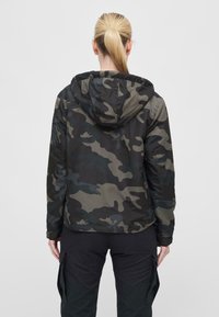 Camouflage jacket in dark green and black with a hood, featuring a textured fabric and fitted design. Visible utility pocket on pants.