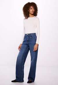 Witte lange mouw top met schouderknopen, gecombineerd met wijde blauwe denim jeans. Soepele stof met een ontspannen pasvorm en subtiele stikseldetails.