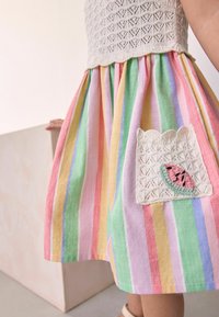 Robe rayée avec un buste en crochet et une poche arborant un motif de pastèque. Couleurs : rose, vert, jaune et bleu. Tissu texturé.