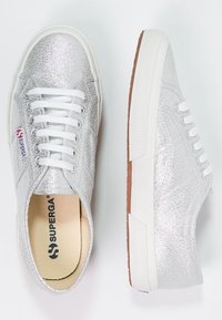 Un paio di sneakers basse argento glitter con lacci bianchi, suole di gomma bianche e etichetta del marchio Superga.