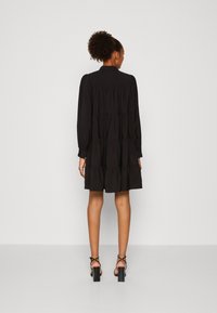 Svart, lager-på-lager klänning med långa ärmar och krage, gjord av mjukt material. Har en lös passform och faller ovanför knäna. Svarta högklackade sandaler.