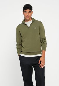 Uomo che indossa un pullover olive green a zip quarti sopra una camicia grigia chiara e pantaloni neri, in piedi con una mano in tasca contro uno sfondo bianco.