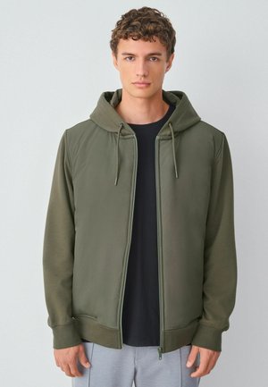 Olivgrön zip-up hoodie med dragsko på huvan, med en mjuk textur och ribbade ärmslut, lagd över en svart skjorta.