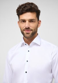 Weißes Hemd mit klassischem Kragen, durchgehender Knopfleiste mit schwarzen Knöpfen und glatter Stoffstruktur. Keine Muster oder Akzente sichtbar.