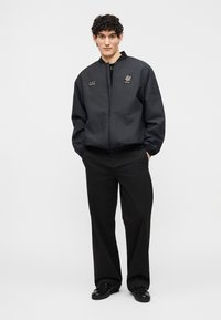 Νεαρός άνδρας που στέκεται με τα χέρια στις τσέπες, φορώντας μαύρο μπουφάν bomber, φαρδιά μαύρη παντελόνια και μαύρα παπούτσια σε λευκό φόντο.