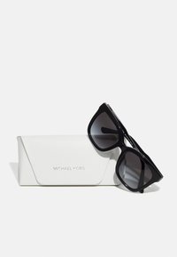 Black square sunglasses with gradient lenses resting on a white, textured case embossed with "Michael Kors" in silver.