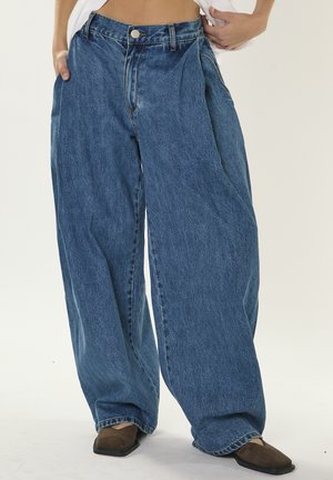 Person iført højtaljede, vidbenede blå jeans med plisseringer, brune sko med firkantet tå og hvid skjorte løftet for at vise linningen.