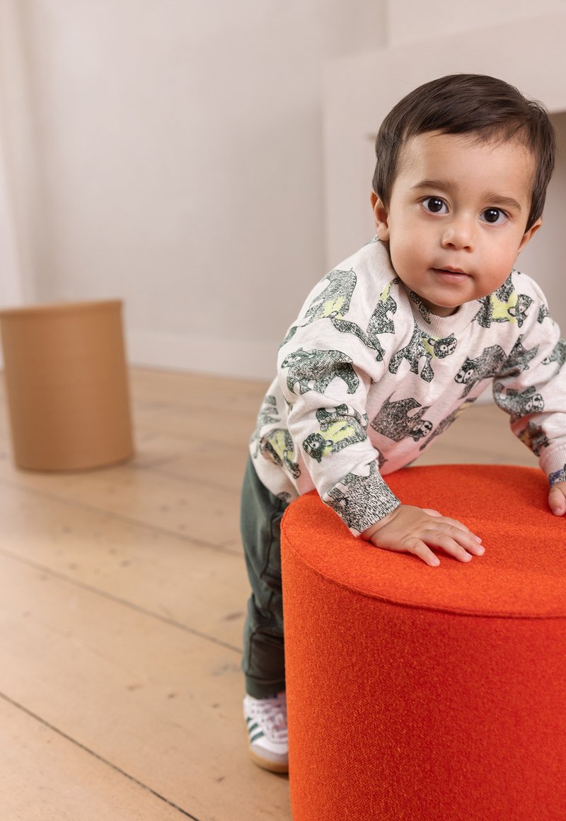 Ljusstolpe i orange med rund form och texturerad tygyta. Barn sträcker ut handen, klädd i en ljus skjorta med djurmönster och mörka byxor.