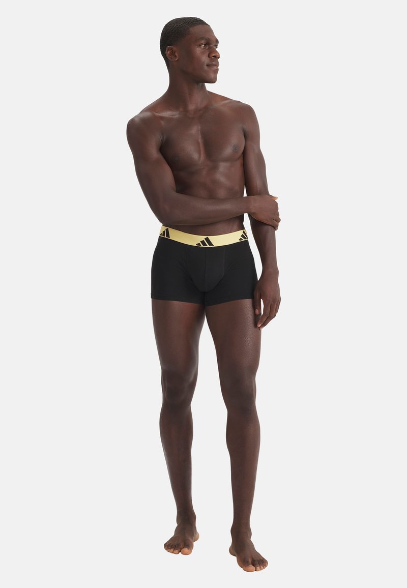 Homme mince à la peau foncée, debout pieds nus, portant un boxer noir avec une large ceinture jaune et logos Adidas, regardant sur le côté.