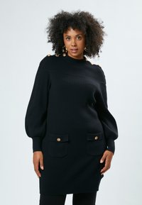 Schwarzes geripptes Strickkleid mit langen Puffärmeln, zwei vorderen Taschen, goldenen Knopfakzenten an den Schultern und einem hohen Kragen.