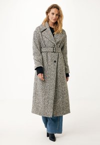 Cappotto in lana con motivo a scacchi in bianco e nero, dotato di vita con cintura, collo a bavero e chiusura con bottoni. Indossato su jeans a gamba larga.