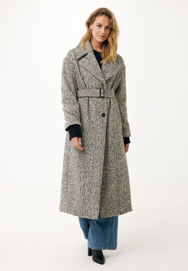 Cappotto in lana con motivo a scacchi in bianco e nero, dotato di vita con cintura, collo a bavero e chiusura con bottoni. Indossato su jeans a gamba larga.