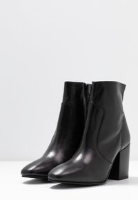 Par de botas de tobillo de cuero negro con punteras puntiagudas, cremalleras laterales y tacones cuadrados sobre una superficie blanca y un fondo gris claro.