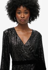 Robe portefeuille en sequins noirs avec un décolleté en V profond, des manches longues et une ceinture en tissu. Texture de surface brillante avec une taille cintrée.