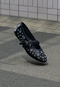 Μαύρο δερμάτινο επίπεδο παπούτσι με κυκλικά ανοίγματα και διακοσμητικά περιλαίμια. Διαθέτει ρυθμιζόμενο διπλό λουράκι και στρογγυλεμένο σχεδιασμό μύτης.