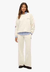 Sweat-shirt blanc surdimensionné avec le logo « Superdry », associé à une chemise à rayures bleues et un pantalon large crème. Chaussures noires et détail de patch de drapeau.