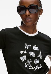 Czarny t-shirt z białymi akcentami, z graficznym nadrukiem akcesoriów, kota i wieży Eiffla w okręgu. Modelka nosi oversize'owe czarne okulary przeciwsłoneczne.