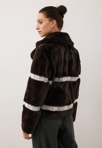 Bruine faux fur-jack met een brede kraag, voorzien van contrasterende witte banden met het label "BIZUU" rond de taille en mouwen.