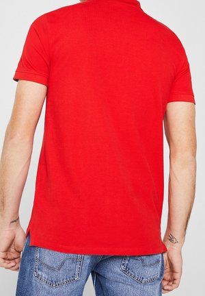 Mand iført en ensfarvet rød poloshirt med korte ærmer og blå jeans, set bagfra, med små tatoveringer på begge underarme.