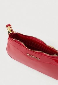 Mala tipo pochete em couro envernizado vermelho com ferragens e fechos dourados, apresentando um detalhe de logótipo na lateral e uma textura lisa e brilhante.