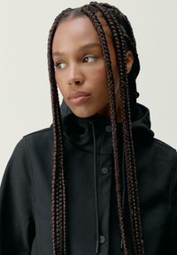 Črna jakna z visokim ovratnikom, ki ima gladko teksturo in gumbne zapore. Pleteni lasje padajo čez ramena.