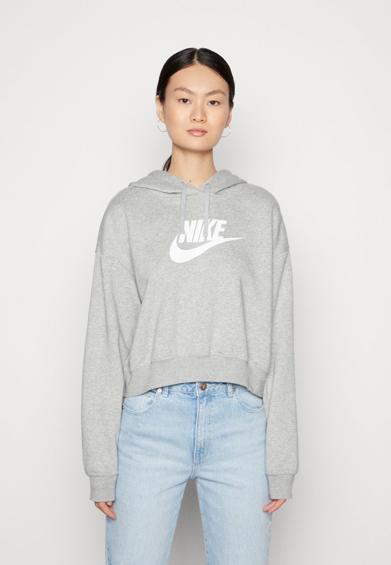 Šedá zkrácená mikina vyrobená z fleecu, s bílým logem Nike, klokaní kapsou a šňůrkou na kapuci. V kombinaci s bleděmodrými džíny.