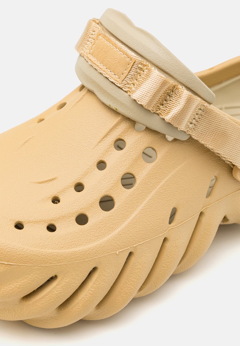 Zoccolo beige realizzato in plastica testurizzata con fori di ventilazione e una leggera tracolla con un accento in tessuto. Suola spessa con scanalature per la trazione.