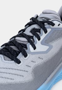 Scarpa sportiva grigio chiaro con lacci scuri, tomaia in mesh, tessuto strutturato e suola blu. Presenta un dettaglio liscio in plastica sul lato.