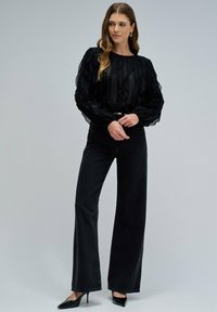 Blusa negra texturizada con mangas con volantes, combinada con unos vaqueros negros de cintura alta. La modelo lleva unos tacones negros puntiagudos y un cinturón sutil.