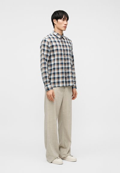 Νεαρός άνδρας όρθιος, φοράει μπλε και καφέ πουκάμισο με κουμπιά καρώ, μπεζ παντελόνι φαρδύ και λευκά αθλητικά παπούτσια, μπροστά σε απλό λευκό φόντο.