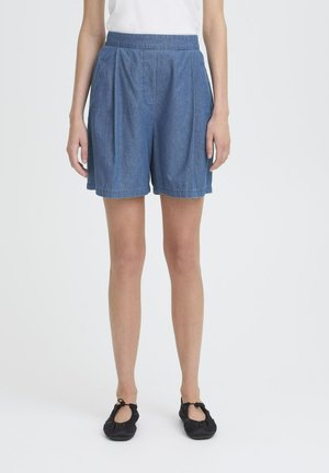 Jeansshorts in mittelblau mit einem gefalteten Design, elastischem Bund und genähten Säumen. Tragen kombiniert mit einem schlichten weißen Oberteil und schwarzen Ballerinas.