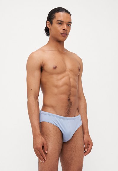 Svetlo modri kopalni boksarji z gladko teksturo in elastičnim pasom, s poudarkom na majhnem logotipu ob strani. Model stoji obrnjen levo.