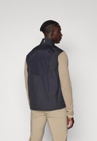 Utenermet navy vest med høy krage, glatt tekstur og baklomme, iført over en beige langermet skjorte.