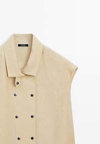 Sleeveless beige shirt with wide collar and two vertical rows of black snap buttons down the front.